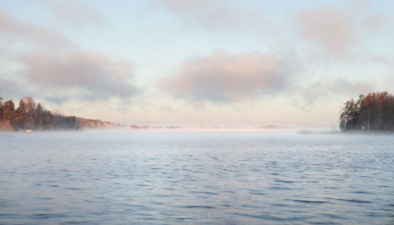 Sea smoke rising from Lake Lohja at sunrise, pale pink sky reflected on water and island visible through mist.